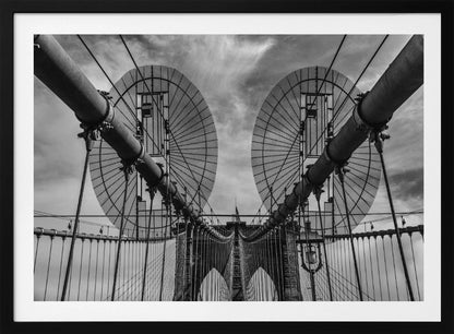 A symmetrical, low-angle black and white photograph of the Brooklyn Bridge's suspension cables and towers against a cloudy sky, framed in silver. Poster
