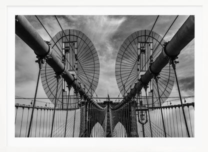 A symmetrical, low-angle black and white photograph of the Brooklyn Bridge's suspension cables and towers against a cloudy sky, framed in silver. Poster