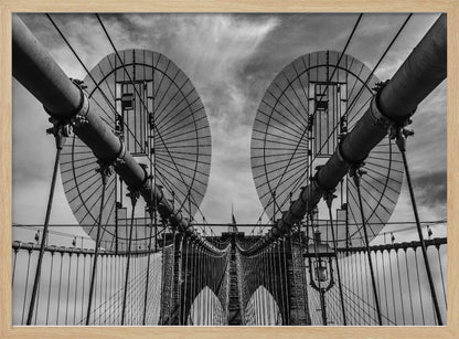 A symmetrical, low-angle black and white photograph of the Brooklyn Bridge's suspension cables and towers against a cloudy sky, framed in silver. Poster