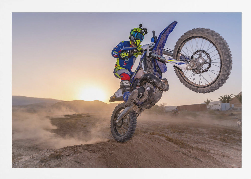 A motocross rider in blue and yellow gear is captured mid-air on a blue dirt bike, performing a jump against a dusty landscape with a setting sun in the background. The image is enclosed in a silver frame. Wall Art