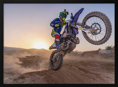 A motocross rider in blue and yellow gear is captured mid-air on a blue dirt bike, performing a jump against a dusty landscape with a setting sun in the background. The image is enclosed in a silver frame. Wall Art