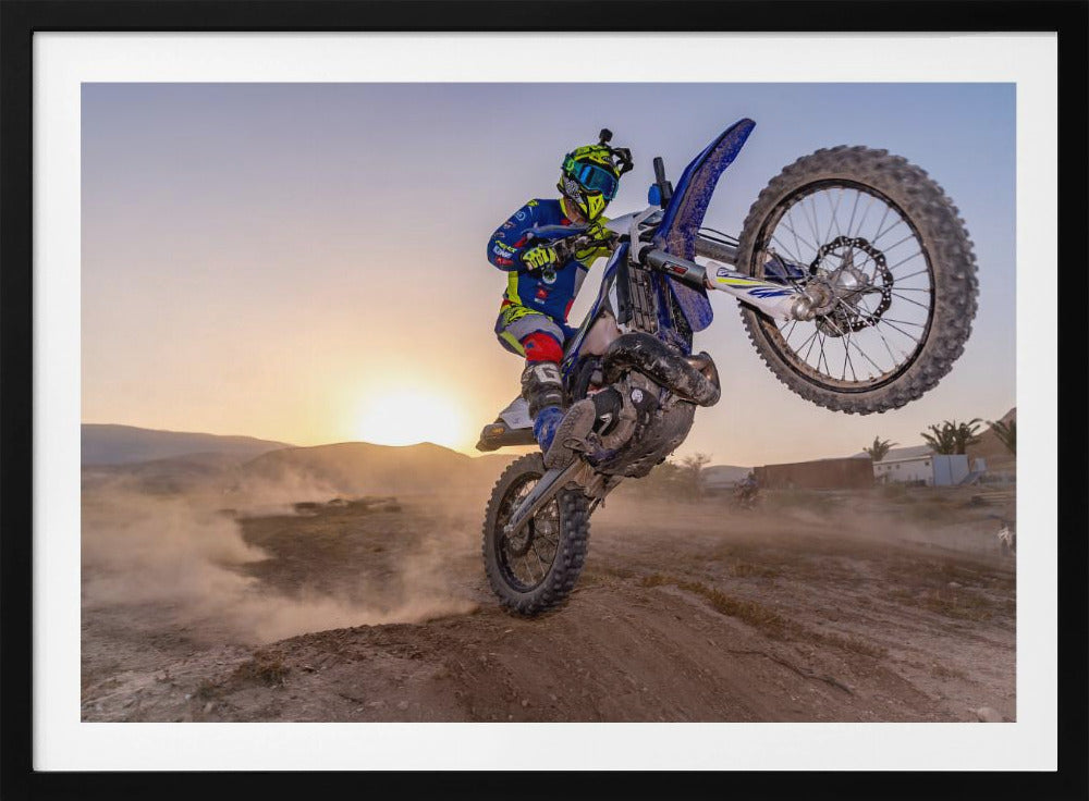 A motocross rider in blue and yellow gear is captured mid-air on a blue dirt bike, performing a jump against a dusty landscape with a setting sun in the background. The image is enclosed in a silver frame. Wall Art