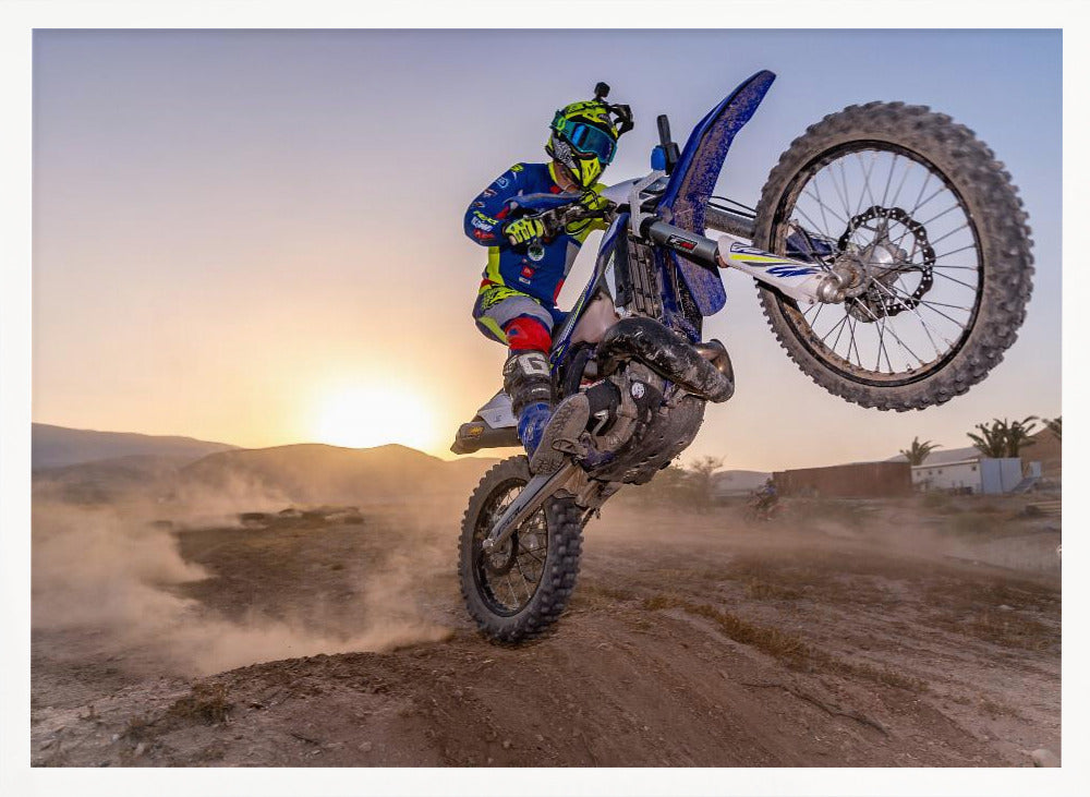 A motocross rider in blue and yellow gear is captured mid-air on a blue dirt bike, performing a jump against a dusty landscape with a setting sun in the background. The image is enclosed in a silver frame. Wall Art
