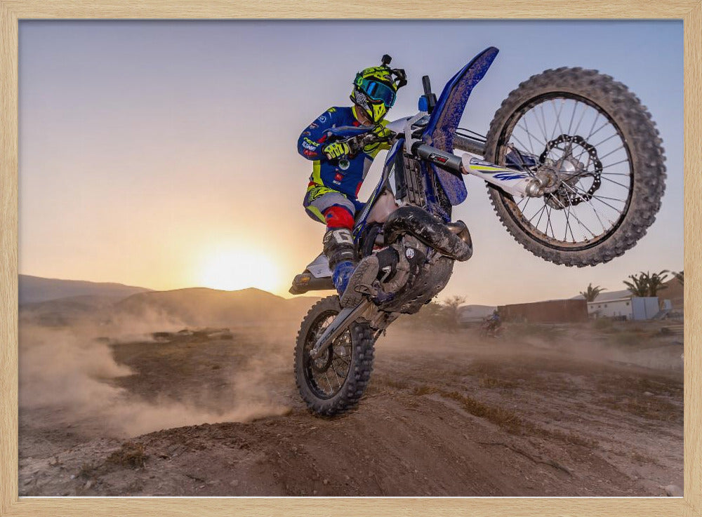 A motocross rider in blue and yellow gear is captured mid-air on a blue dirt bike, performing a jump against a dusty landscape with a setting sun in the background. The image is enclosed in a silver frame. Wall Art