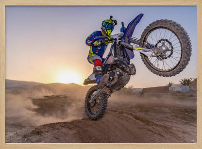 A motocross rider in blue and yellow gear is captured mid-air on a blue dirt bike, performing a jump against a dusty landscape with a setting sun in the background. The image is enclosed in a silver frame. Wall Art