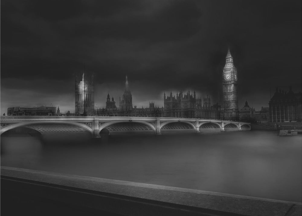 A moody, atmospheric black and white photograph of the Houses of Parliament and Big Ben in London, viewed from across the River Thames. The iconic buildings are softly illuminated against a dark, dramatic sky, with Westminster Bridge arching over the dark, smooth water in the foreground. The image is presented within a silver-colored frame. Artwork