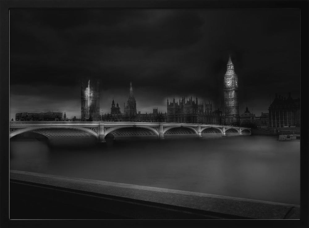 A moody, atmospheric black and white photograph of the Houses of Parliament and Big Ben in London, viewed from across the River Thames. The iconic buildings are softly illuminated against a dark, dramatic sky, with Westminster Bridge arching over the dark, smooth water in the foreground. The image is presented within a silver-colored frame. Artwork
