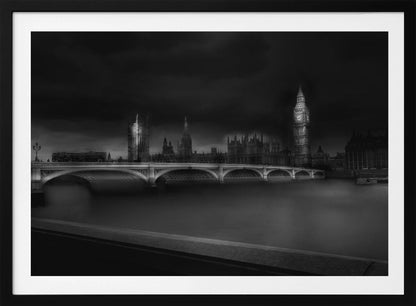 A moody, atmospheric black and white photograph of the Houses of Parliament and Big Ben in London, viewed from across the River Thames. The iconic buildings are softly illuminated against a dark, dramatic sky, with Westminster Bridge arching over the dark, smooth water in the foreground. The image is presented within a silver-colored frame. Artwork