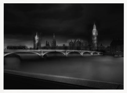 A moody, atmospheric black and white photograph of the Houses of Parliament and Big Ben in London, viewed from across the River Thames. The iconic buildings are softly illuminated against a dark, dramatic sky, with Westminster Bridge arching over the dark, smooth water in the foreground. The image is presented within a silver-colored frame. Artwork