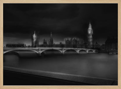 A moody, atmospheric black and white photograph of the Houses of Parliament and Big Ben in London, viewed from across the River Thames. The iconic buildings are softly illuminated against a dark, dramatic sky, with Westminster Bridge arching over the dark, smooth water in the foreground. The image is presented within a silver-colored frame. Artwork