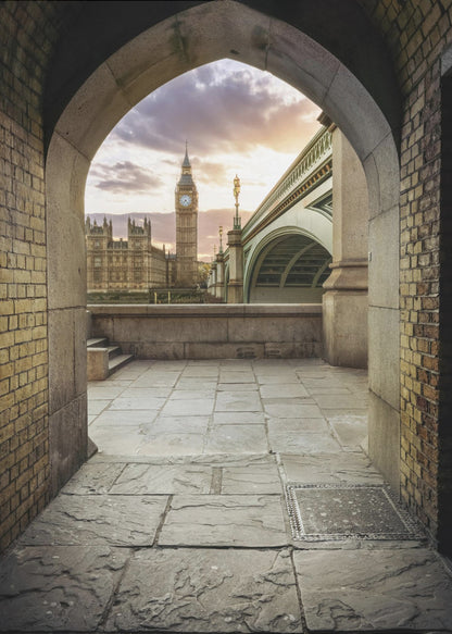 A scenic view of London's Big Ben clock tower and the Houses of Parliament as seen through a weathered stone archway. The foreground features a stone-paved walkway, and the background shows the iconic landmarks against a cloudy sunset sky with hues of purple and orange. Poster