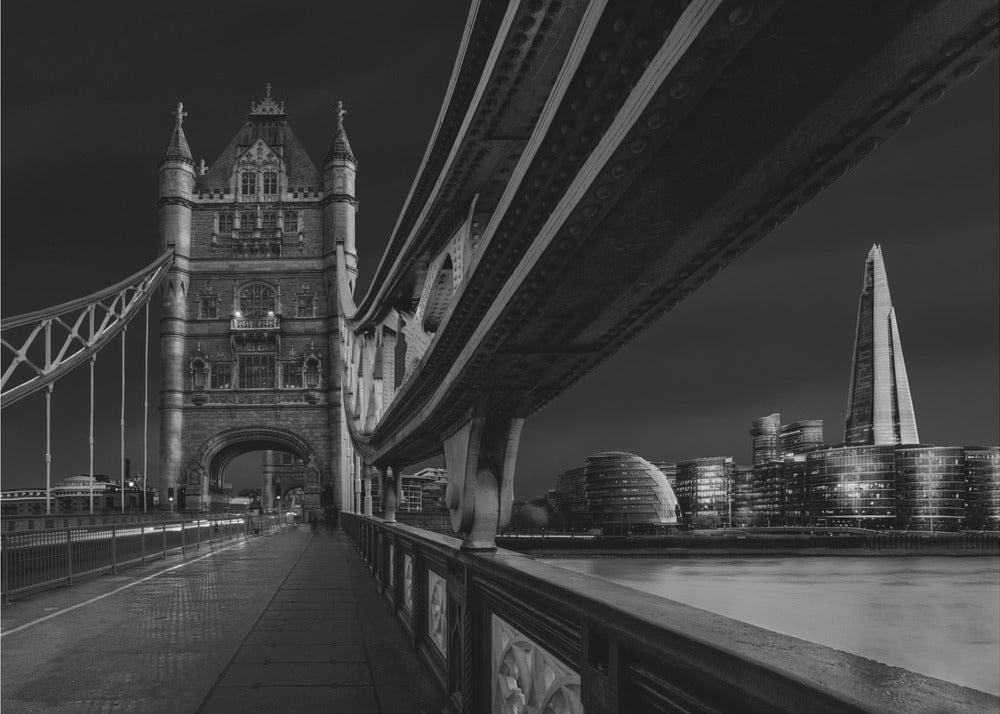 A dramatic, low-angle black and white photograph of London's Tower Bridge at night, taken from the pedestrian walkway. The image showcases the architectural details of the bridge and the illuminated modern city skyline, including The Shard, across the River Thames. Print