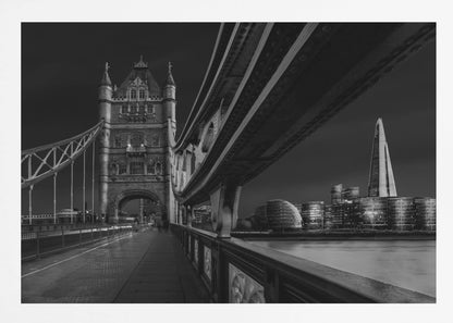A dramatic, low-angle black and white photograph of London's Tower Bridge at night, taken from the pedestrian walkway. The image showcases the architectural details of the bridge and the illuminated modern city skyline, including The Shard, across the River Thames. Print