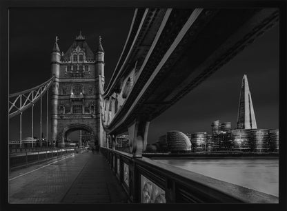 A dramatic, low-angle black and white photograph of London's Tower Bridge at night, taken from the pedestrian walkway. The image showcases the architectural details of the bridge and the illuminated modern city skyline, including The Shard, across the River Thames. Print