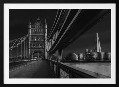 A dramatic, low-angle black and white photograph of London's Tower Bridge at night, taken from the pedestrian walkway. The image showcases the architectural details of the bridge and the illuminated modern city skyline, including The Shard, across the River Thames. Print