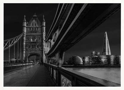 A dramatic, low-angle black and white photograph of London's Tower Bridge at night, taken from the pedestrian walkway. The image showcases the architectural details of the bridge and the illuminated modern city skyline, including The Shard, across the River Thames. Print
