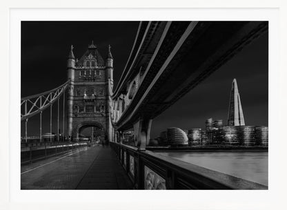 A dramatic, low-angle black and white photograph of London's Tower Bridge at night, taken from the pedestrian walkway. The image showcases the architectural details of the bridge and the illuminated modern city skyline, including The Shard, across the River Thames. Print