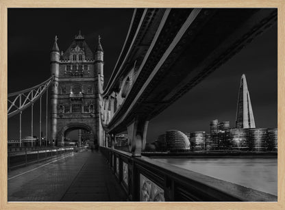 A dramatic, low-angle black and white photograph of London's Tower Bridge at night, taken from the pedestrian walkway. The image showcases the architectural details of the bridge and the illuminated modern city skyline, including The Shard, across the River Thames. Print