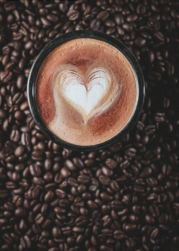 A top-down view of a cappuccino in a black mug with a white heart-shaped foam art design, placed on a bed of dark roasted coffee beans. Artwork