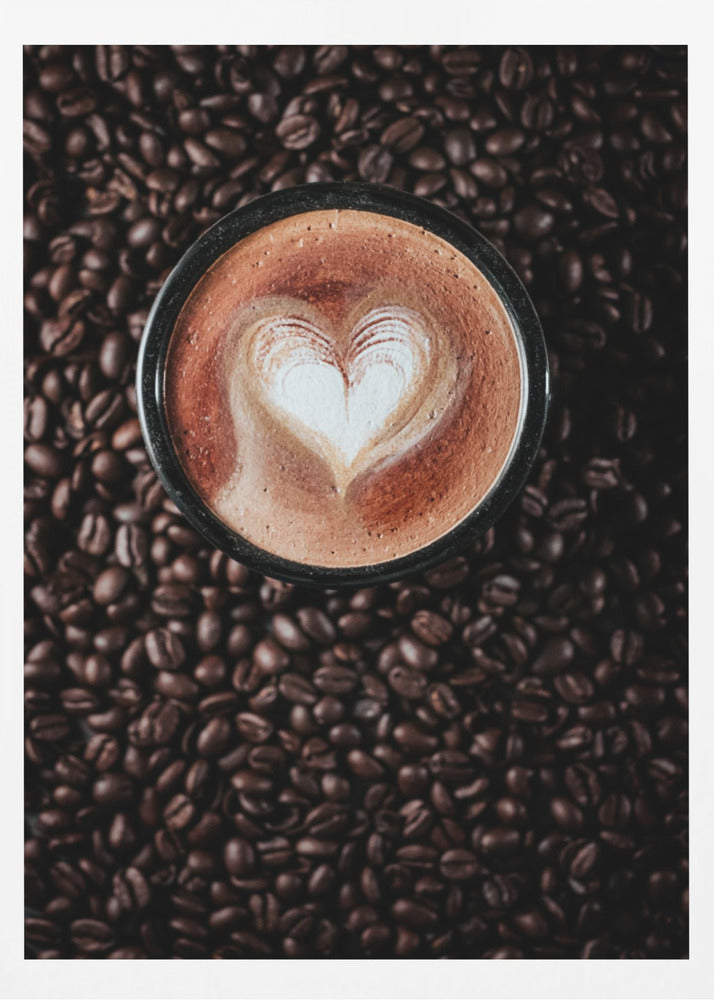 A top-down view of a cappuccino in a black mug with a white heart-shaped foam art design, placed on a bed of dark roasted coffee beans. Artwork