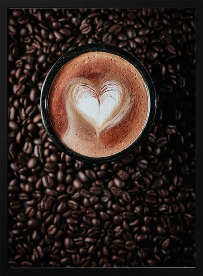 A top-down view of a cappuccino in a black mug with a white heart-shaped foam art design, placed on a bed of dark roasted coffee beans. Artwork