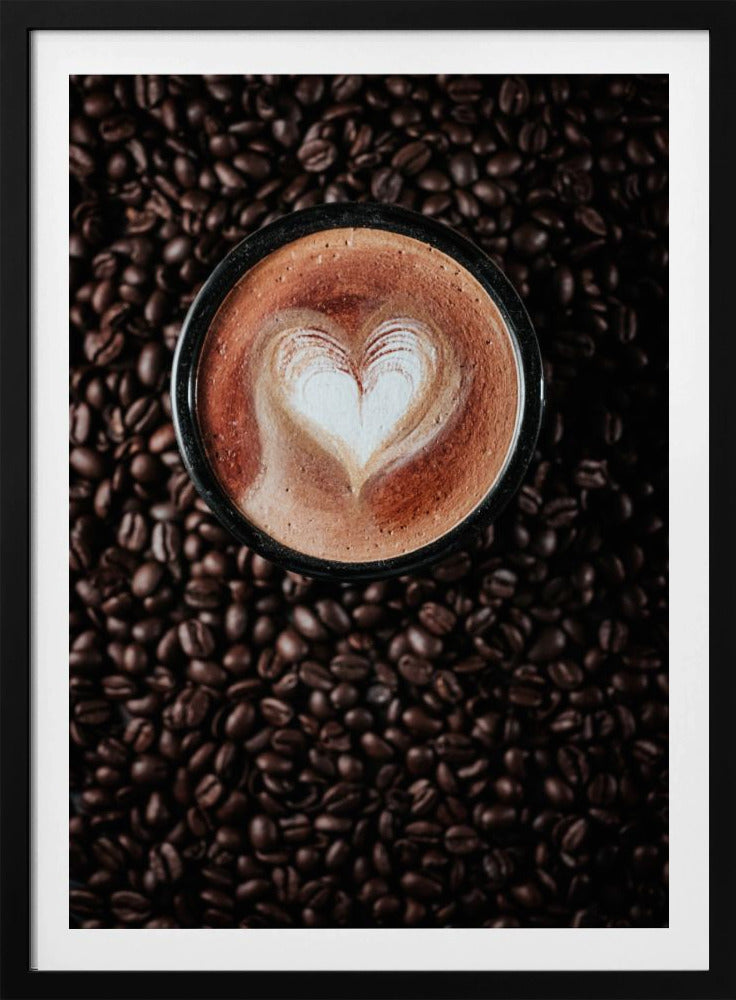 A top-down view of a cappuccino in a black mug with a white heart-shaped foam art design, placed on a bed of dark roasted coffee beans. Artwork