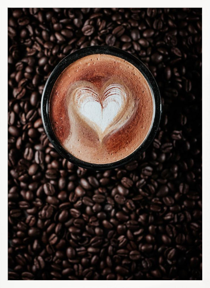 A top-down view of a cappuccino in a black mug with a white heart-shaped foam art design, placed on a bed of dark roasted coffee beans. Artwork