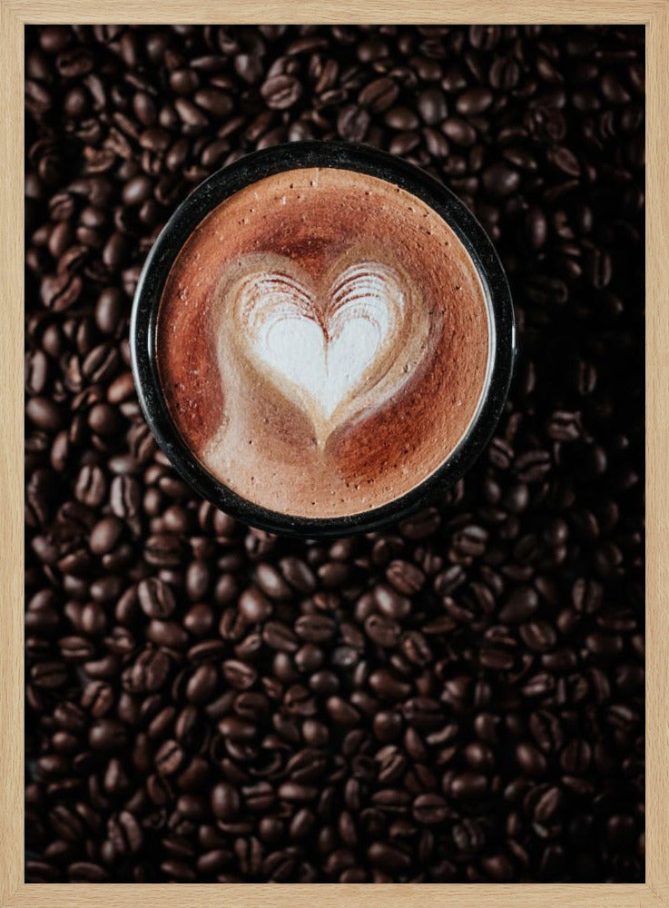 A top-down view of a cappuccino in a black mug with a white heart-shaped foam art design, placed on a bed of dark roasted coffee beans. Artwork