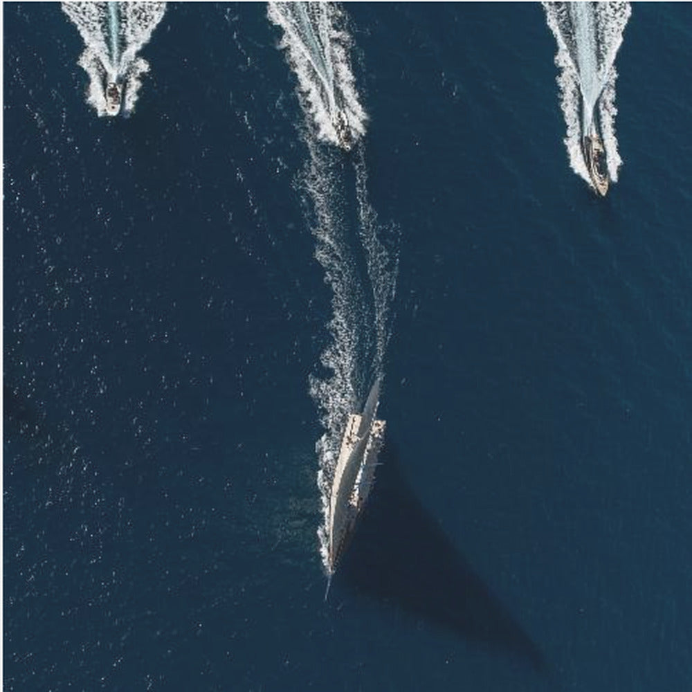 An aerial photograph from directly above shows a large sailboat and three smaller motorboats moving across a deep blue sea, leaving white wakes behind them. The sailboat is in the foreground, casting a long shadow, while the smaller boats are positioned further back. The entire image is enclosed in a light-colored wooden frame. Wall Art