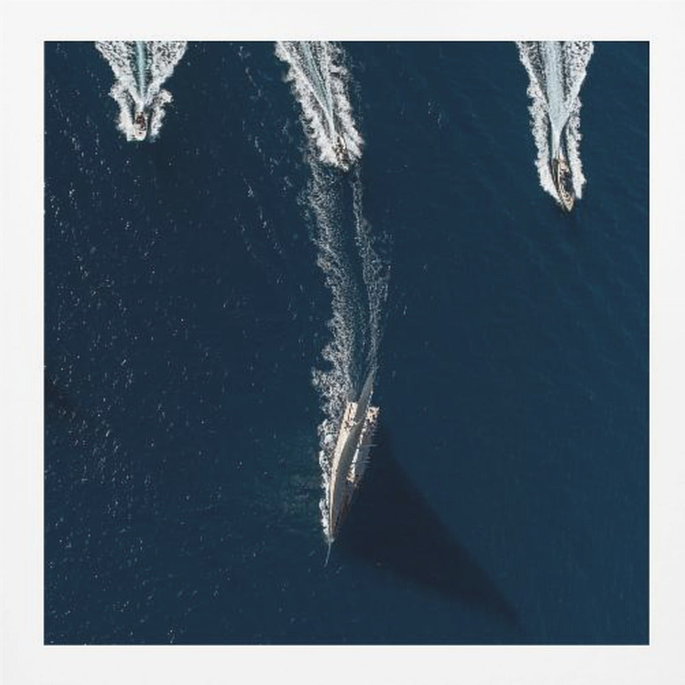 An aerial photograph from directly above shows a large sailboat and three smaller motorboats moving across a deep blue sea, leaving white wakes behind them. The sailboat is in the foreground, casting a long shadow, while the smaller boats are positioned further back. The entire image is enclosed in a light-colored wooden frame. Wall Art