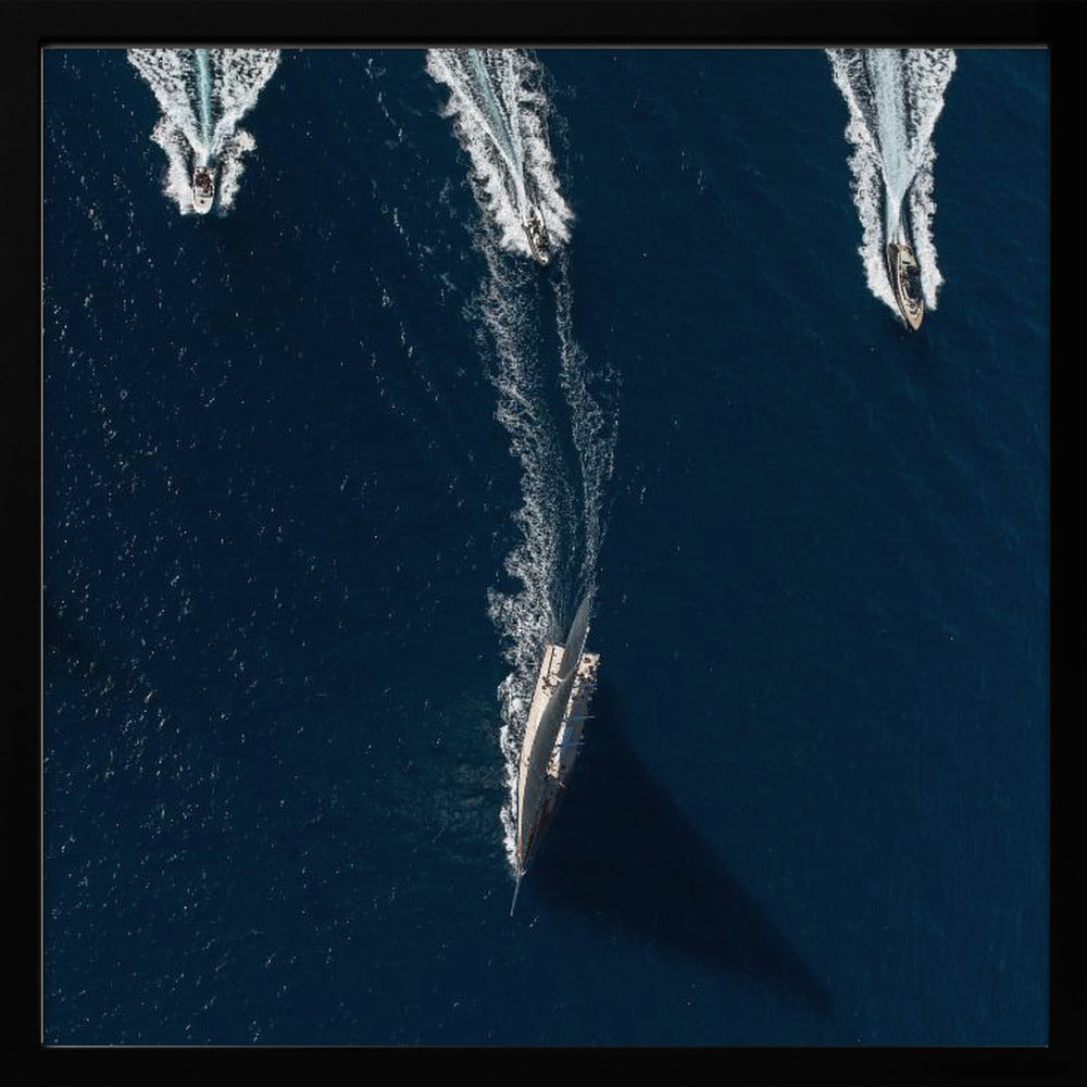 An aerial photograph from directly above shows a large sailboat and three smaller motorboats moving across a deep blue sea, leaving white wakes behind them. The sailboat is in the foreground, casting a long shadow, while the smaller boats are positioned further back. The entire image is enclosed in a light-colored wooden frame. Wall Art