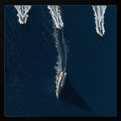 An aerial photograph from directly above shows a large sailboat and three smaller motorboats moving across a deep blue sea, leaving white wakes behind them. The sailboat is in the foreground, casting a long shadow, while the smaller boats are positioned further back. The entire image is enclosed in a light-colored wooden frame. Wall Art
