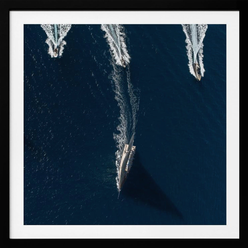 An aerial photograph from directly above shows a large sailboat and three smaller motorboats moving across a deep blue sea, leaving white wakes behind them. The sailboat is in the foreground, casting a long shadow, while the smaller boats are positioned further back. The entire image is enclosed in a light-colored wooden frame. Wall Art
