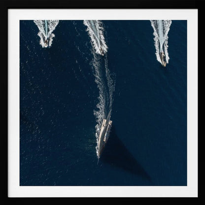 An aerial photograph from directly above shows a large sailboat and three smaller motorboats moving across a deep blue sea, leaving white wakes behind them. The sailboat is in the foreground, casting a long shadow, while the smaller boats are positioned further back. The entire image is enclosed in a light-colored wooden frame. Wall Art
