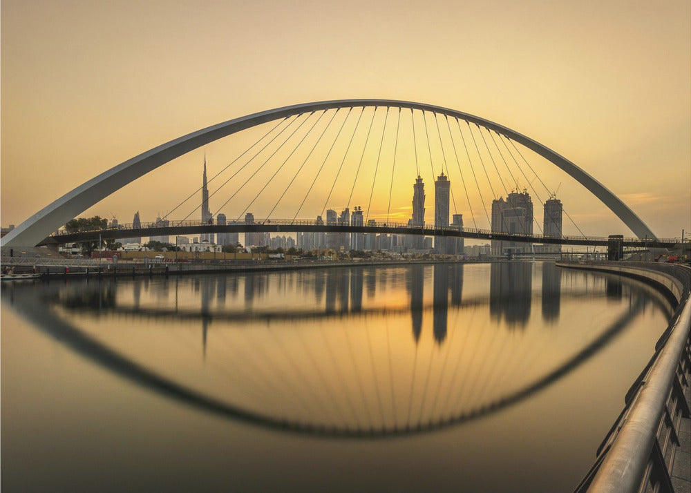 A sweeping view of the Dubai Water Canal Bridge at sunset, its white arch and cables stretching over the calm water. The city skyline, including the Burj Khalifa, is silhouetted against the golden sky, with the entire scene creating a perfect reflection in the water below. Print