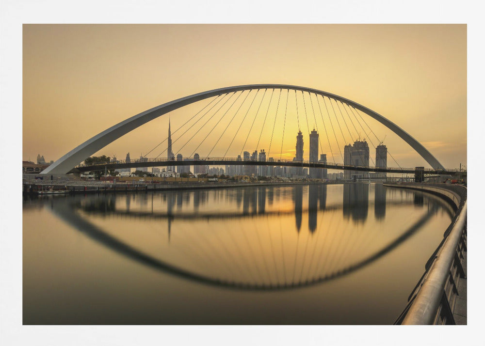 A sweeping view of the Dubai Water Canal Bridge at sunset, its white arch and cables stretching over the calm water. The city skyline, including the Burj Khalifa, is silhouetted against the golden sky, with the entire scene creating a perfect reflection in the water below. Print