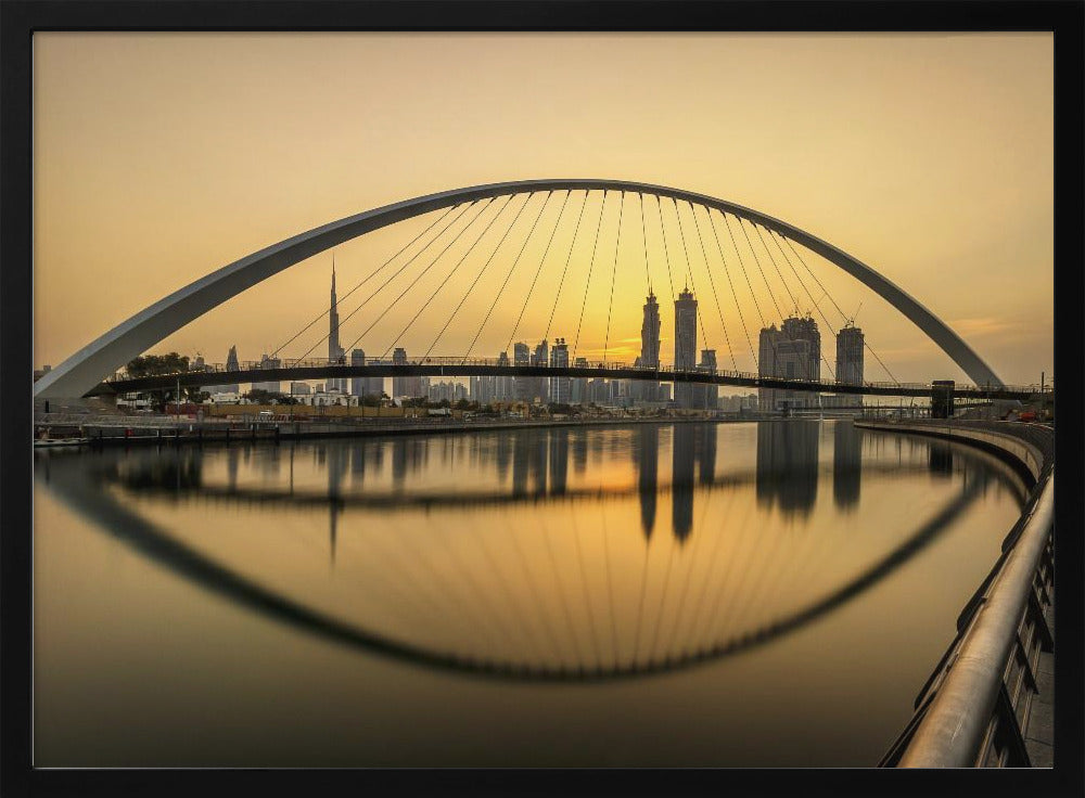 A sweeping view of the Dubai Water Canal Bridge at sunset, its white arch and cables stretching over the calm water. The city skyline, including the Burj Khalifa, is silhouetted against the golden sky, with the entire scene creating a perfect reflection in the water below. Print