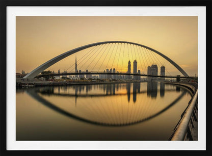 A sweeping view of the Dubai Water Canal Bridge at sunset, its white arch and cables stretching over the calm water. The city skyline, including the Burj Khalifa, is silhouetted against the golden sky, with the entire scene creating a perfect reflection in the water below. Print