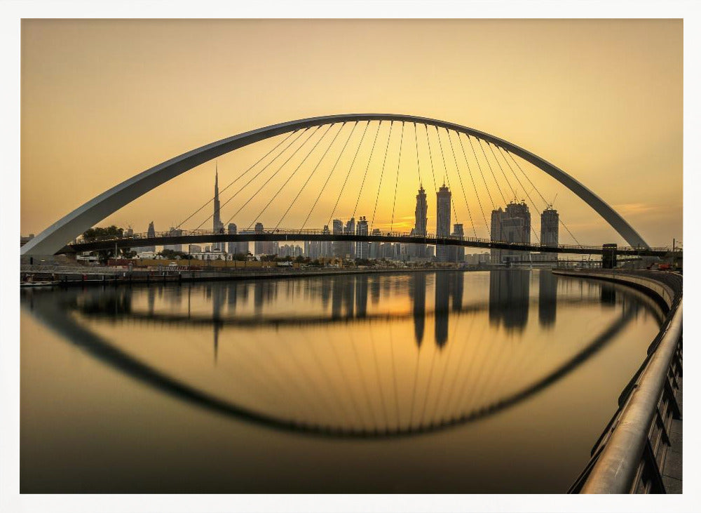 A sweeping view of the Dubai Water Canal Bridge at sunset, its white arch and cables stretching over the calm water. The city skyline, including the Burj Khalifa, is silhouetted against the golden sky, with the entire scene creating a perfect reflection in the water below. Print