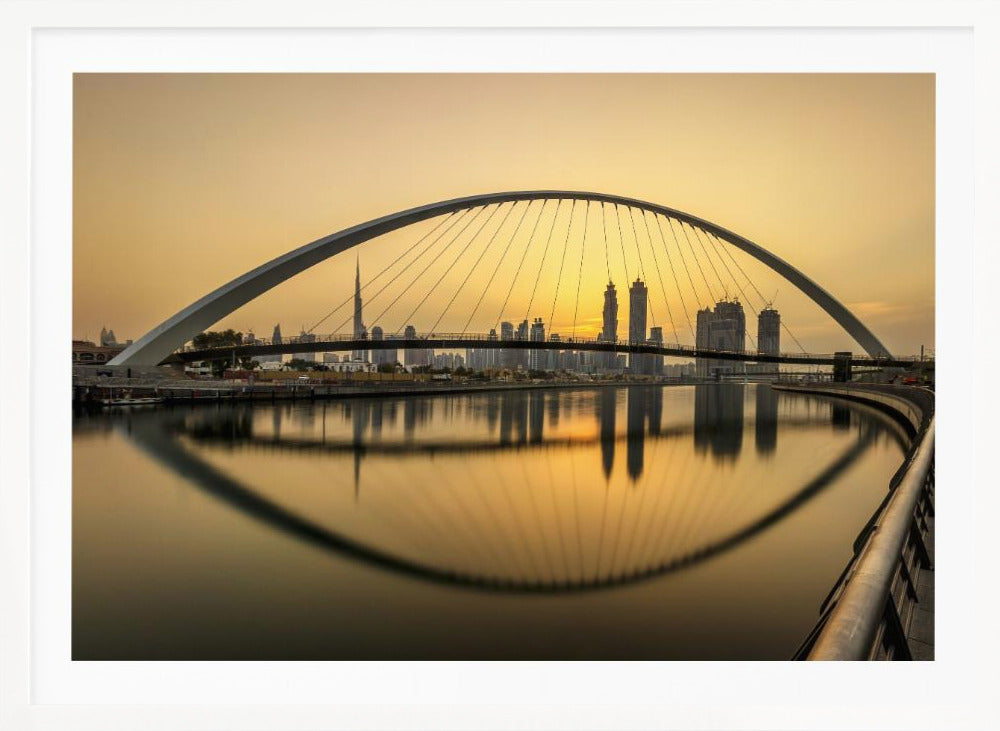A sweeping view of the Dubai Water Canal Bridge at sunset, its white arch and cables stretching over the calm water. The city skyline, including the Burj Khalifa, is silhouetted against the golden sky, with the entire scene creating a perfect reflection in the water below. Print