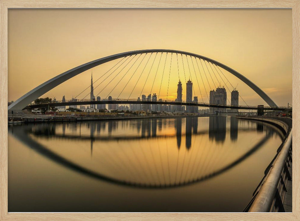 A sweeping view of the Dubai Water Canal Bridge at sunset, its white arch and cables stretching over the calm water. The city skyline, including the Burj Khalifa, is silhouetted against the golden sky, with the entire scene creating a perfect reflection in the water below. Print