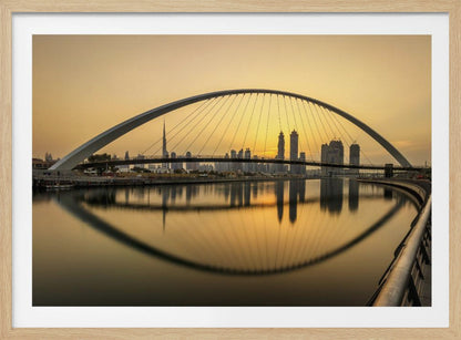 A sweeping view of the Dubai Water Canal Bridge at sunset, its white arch and cables stretching over the calm water. The city skyline, including the Burj Khalifa, is silhouetted against the golden sky, with the entire scene creating a perfect reflection in the water below. Print