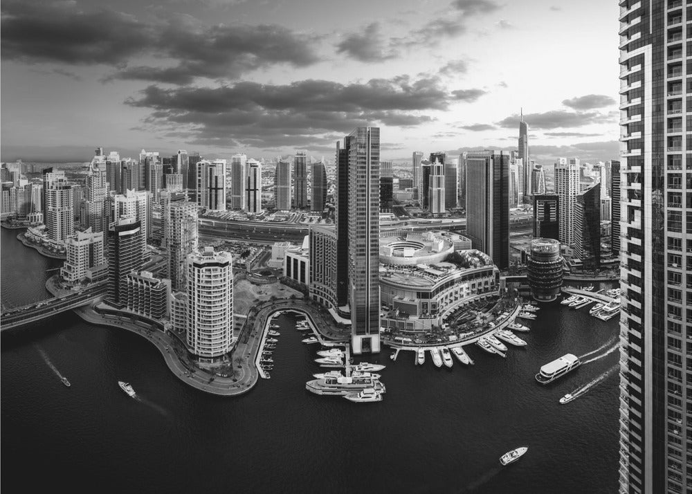 A framed, black and white panoramic photograph of the Dubai Marina taken from a high vantage point. The image captures the dense cityscape with numerous modern skyscrapers surrounding a winding waterway filled with docked yachts and moving boats. The sky is filled with dramatic, dark clouds, adding to the moody atmosphere of the urban landscape. Decor