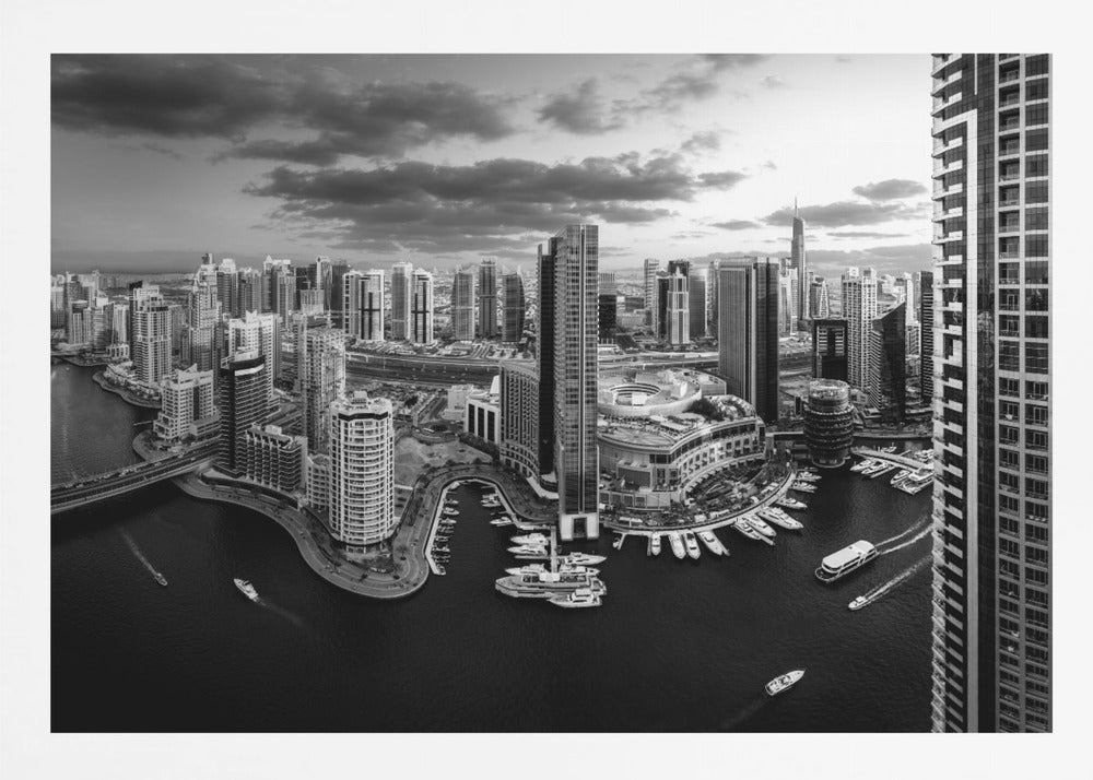 A framed, black and white panoramic photograph of the Dubai Marina taken from a high vantage point. The image captures the dense cityscape with numerous modern skyscrapers surrounding a winding waterway filled with docked yachts and moving boats. The sky is filled with dramatic, dark clouds, adding to the moody atmosphere of the urban landscape. Decor