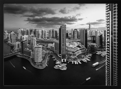 A framed, black and white panoramic photograph of the Dubai Marina taken from a high vantage point. The image captures the dense cityscape with numerous modern skyscrapers surrounding a winding waterway filled with docked yachts and moving boats. The sky is filled with dramatic, dark clouds, adding to the moody atmosphere of the urban landscape. Decor