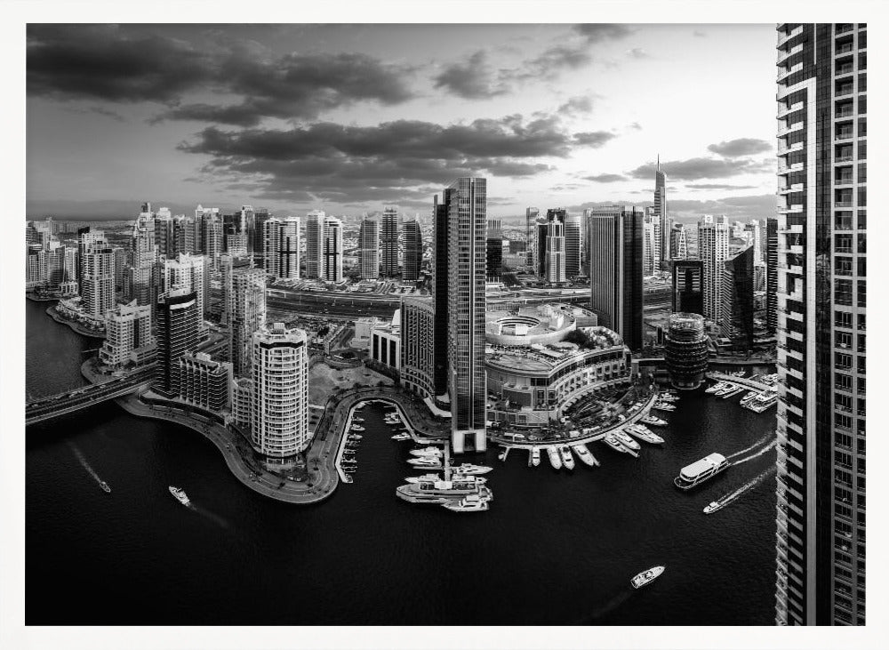 A framed, black and white panoramic photograph of the Dubai Marina taken from a high vantage point. The image captures the dense cityscape with numerous modern skyscrapers surrounding a winding waterway filled with docked yachts and moving boats. The sky is filled with dramatic, dark clouds, adding to the moody atmosphere of the urban landscape. Decor