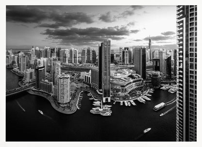 A framed, black and white panoramic photograph of the Dubai Marina taken from a high vantage point. The image captures the dense cityscape with numerous modern skyscrapers surrounding a winding waterway filled with docked yachts and moving boats. The sky is filled with dramatic, dark clouds, adding to the moody atmosphere of the urban landscape. Decor