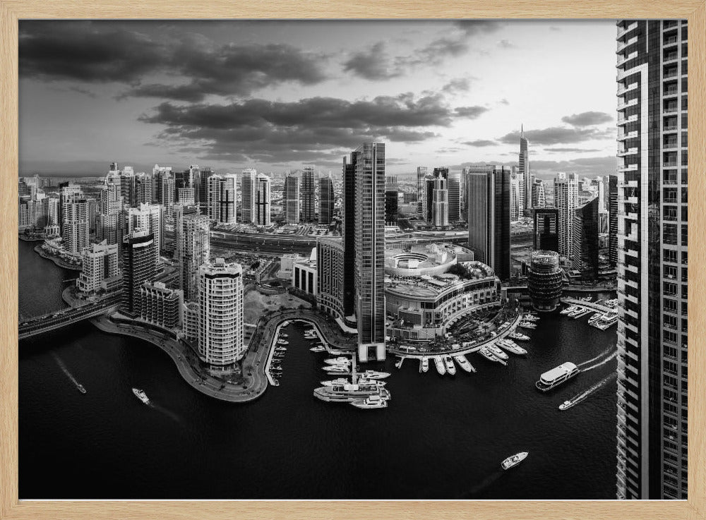 A framed, black and white panoramic photograph of the Dubai Marina taken from a high vantage point. The image captures the dense cityscape with numerous modern skyscrapers surrounding a winding waterway filled with docked yachts and moving boats. The sky is filled with dramatic, dark clouds, adding to the moody atmosphere of the urban landscape. Decor