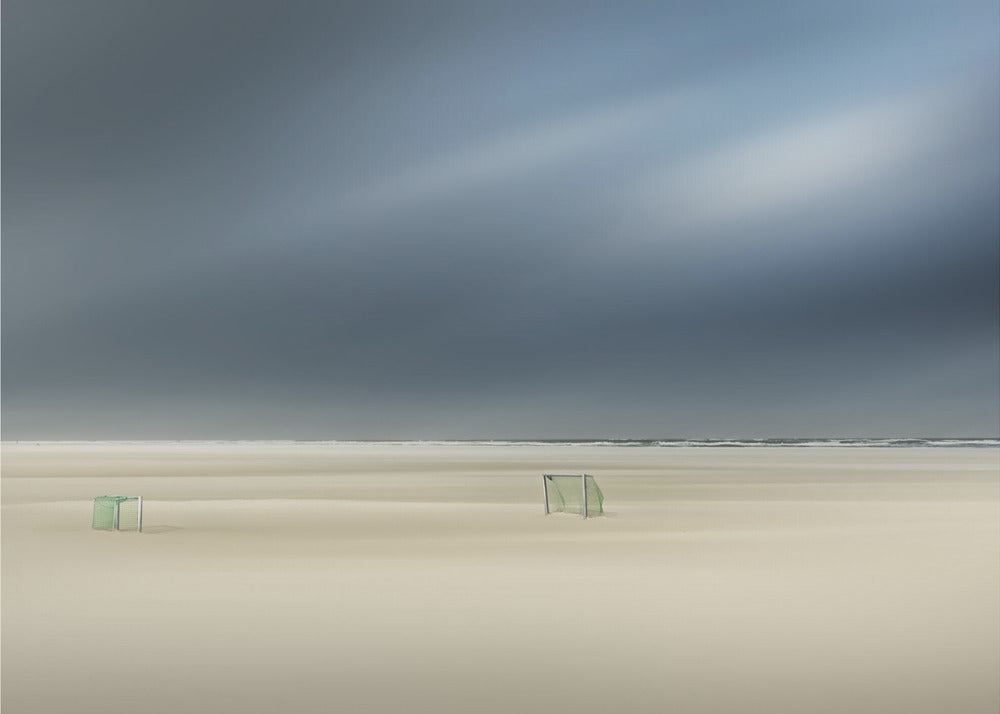 A minimalist long-exposure photograph of a vast, empty beach under a dark, moody sky. Two small, solitary soccer goals sit on the pale sand, with the ocean visible as a thin line in the distance. The artwork is presented in a silver frame. Artwork