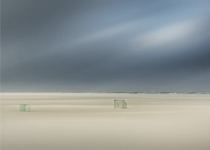 A minimalist long-exposure photograph of a vast, empty beach under a dark, moody sky. Two small, solitary soccer goals sit on the pale sand, with the ocean visible as a thin line in the distance. The artwork is presented in a silver frame. Artwork